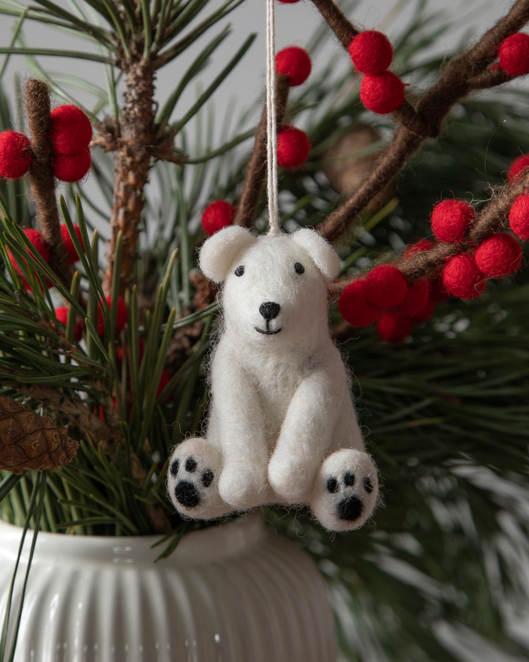 Filzanhänger "sitzender Eisbär" von Gray & Sif aus handgefertigtem weißen Filz. Der Eisbär ist an einem grünen Kiefernzweig befestigt. Weitere Dekoäste aus Filz mit roten Beeren daran sind  in die gleiche Vase gestellt, wie der Kiefernzweig.