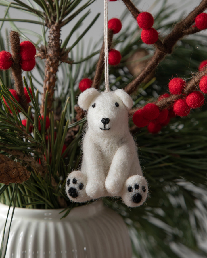 Filzanhänger "sitzender Eisbär" von Gray & Sif aus handgefertigtem weißen Filz. Der Eisbär ist an einem grünen Kiefernzweig befestigt. Weitere Dekoäste aus Filz mit roten Beeren daran sind  in die gleiche Vase gestellt, wie der Kiefernzweig.