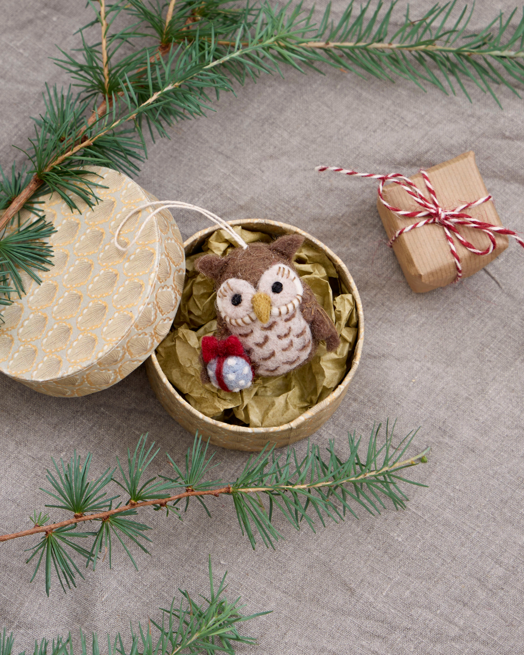 Weihnachtlicher Filzanhänger "Eule mit Geschenk" selbst als kleines Geschenk verpackt in einer runden Dose mit goldenem Papier und einem weiteren kleinen Geschenk daneben.