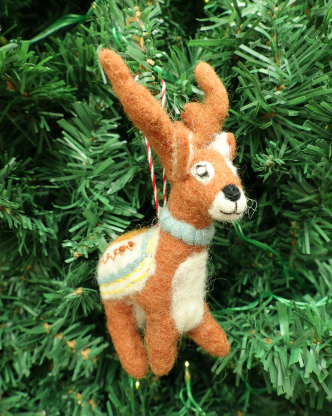 Hirsch Filzanhänger "Noah" von Grace und Gloria hängt am Tannenbaum. Der Hirsch trägt ein hellblaues Halsband und ein weiß/blaues Deckchen.
