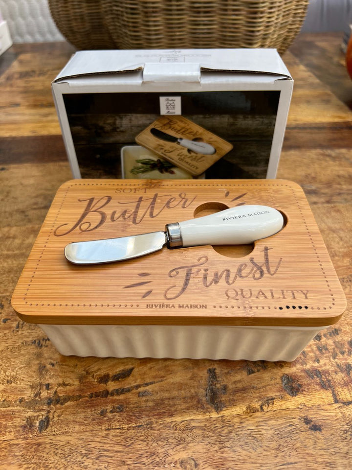 Butterdose mit kleinem Messer „Finest Quality Butter Dish“ von Rivièra Maison auf einem rustikalen Holztisch, mit Holzdeckel und aufgelegtem Buttermesser, Verpackung im Hintergrund sichtbar.