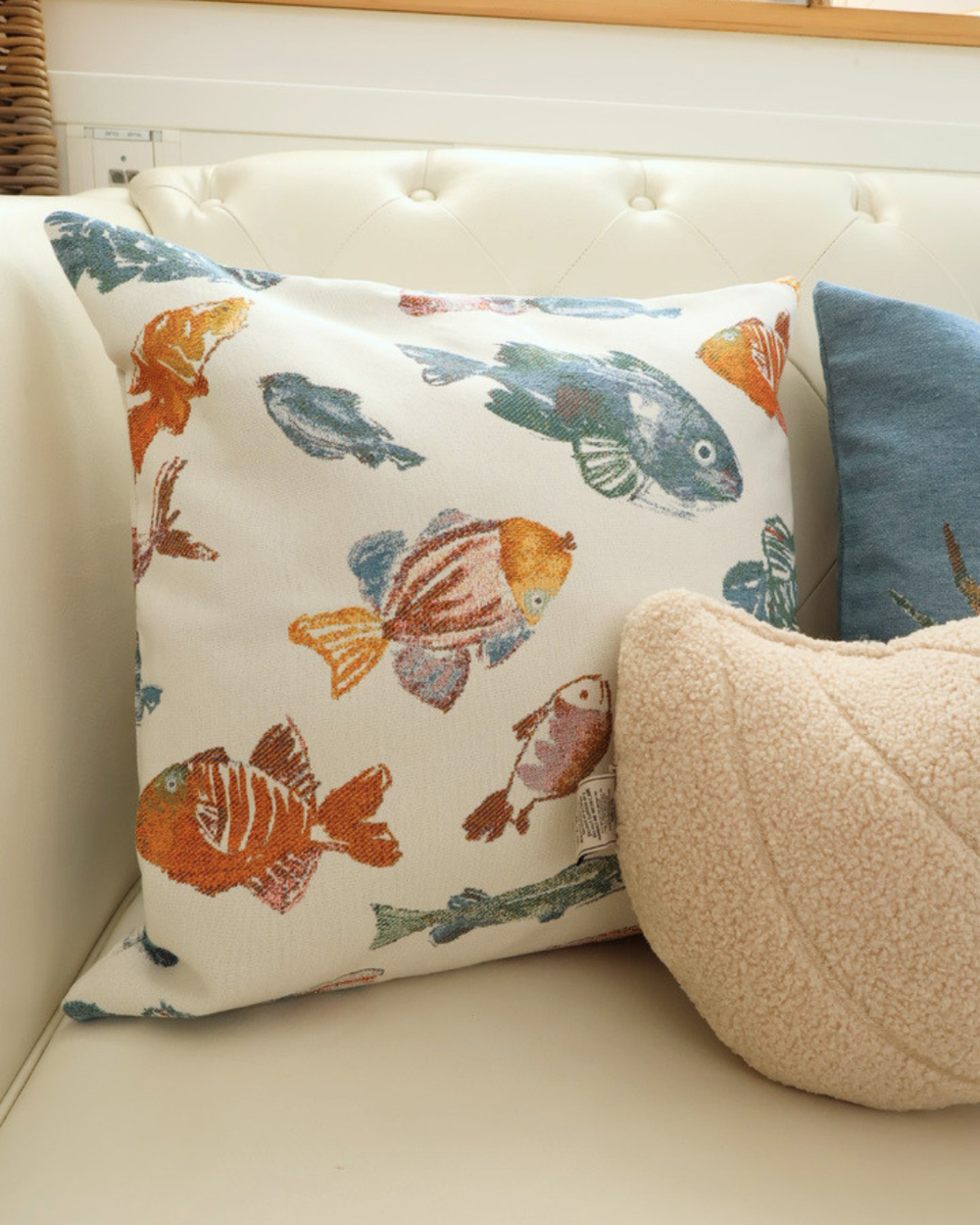 Maritimes Fisch Kissen "Glup" von Sander Table Home. Das Motiv besteht aus einem weißen Hintergrund und verschiedenen Fischen im Vordergrund. Das Kissen liegt auf einem weißen Sofa neben 2 anderen Kissen.