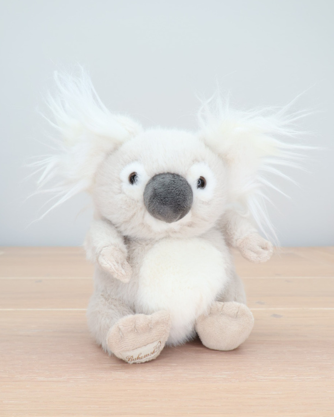 Ein cremefarbener Baby Koala von Bukowski sitzt aufrecht auf einem hellen Holzboden. Das Kuscheltier hat große, schwarze Augen und flauschige Ohren.