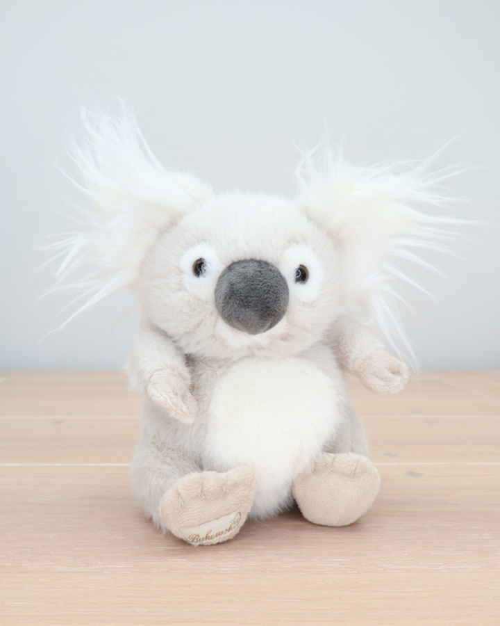 Ein cremefarbener Baby Koala von Bukowski sitzt aufrecht auf einem hellen Holzboden. Das Kuscheltier hat große, schwarze Augen und flauschige Ohren.