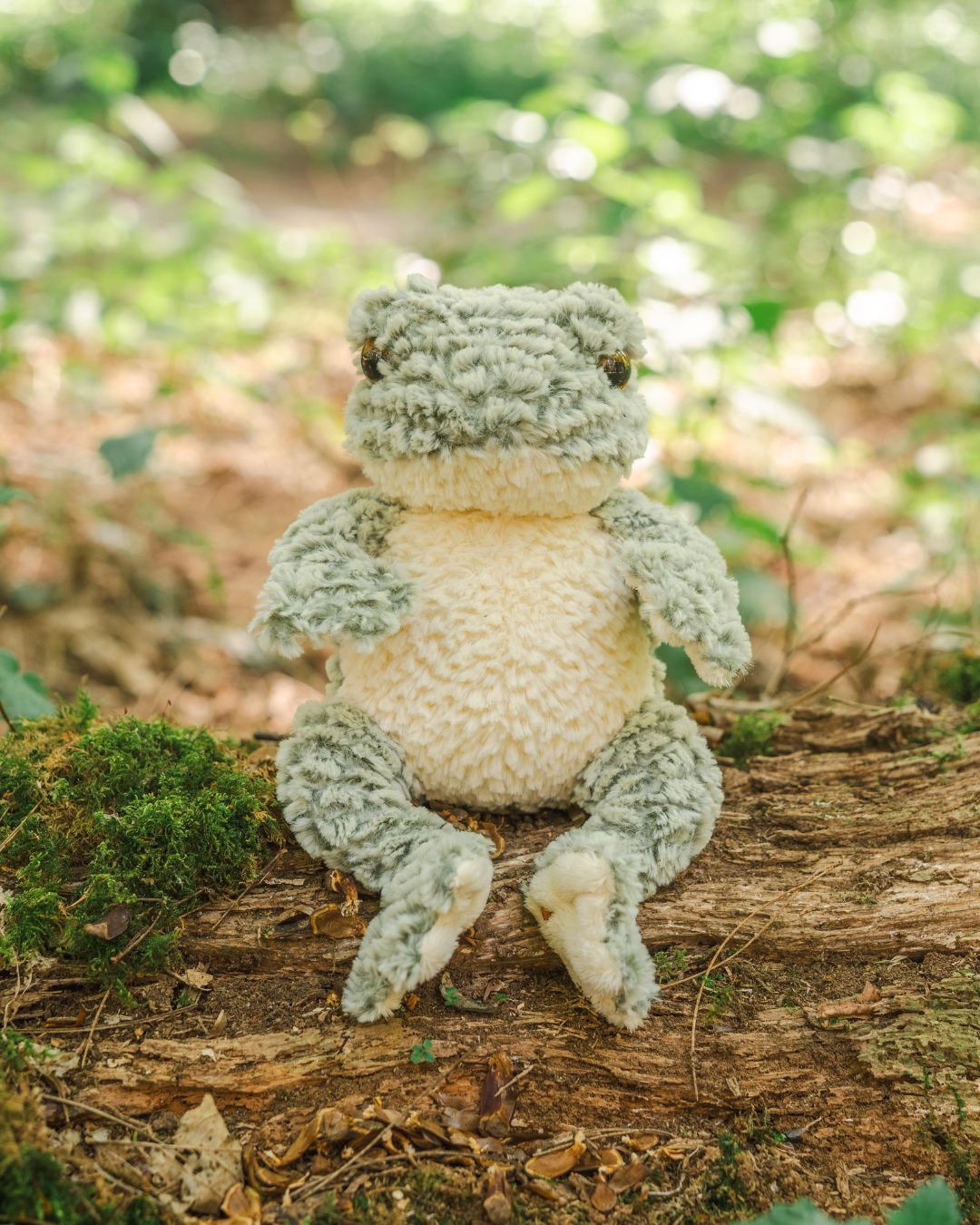 Frosch Kuscheltier „Elliott“ von Wrendale Designs sitzend auf einem Baumstamm im Wald, dekorativ inszeniert in natürlicher Umgebung.