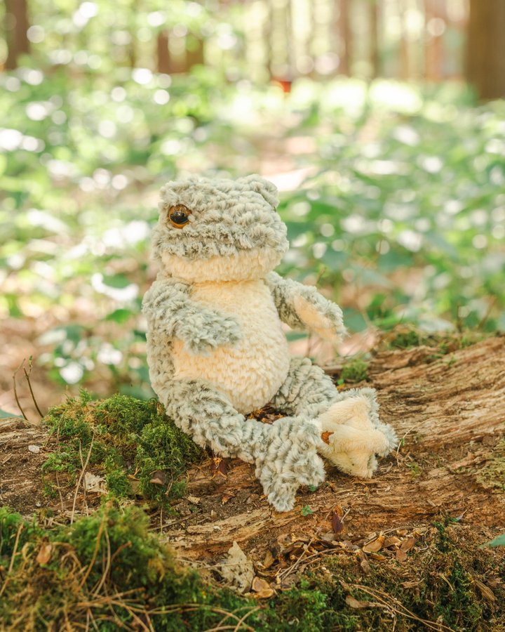 Seitlich fotografiertes Frosch Kuscheltier „Elliott“ von Wrendale Designs auf einem Baumstamm, mit Blick in die Waldszene.