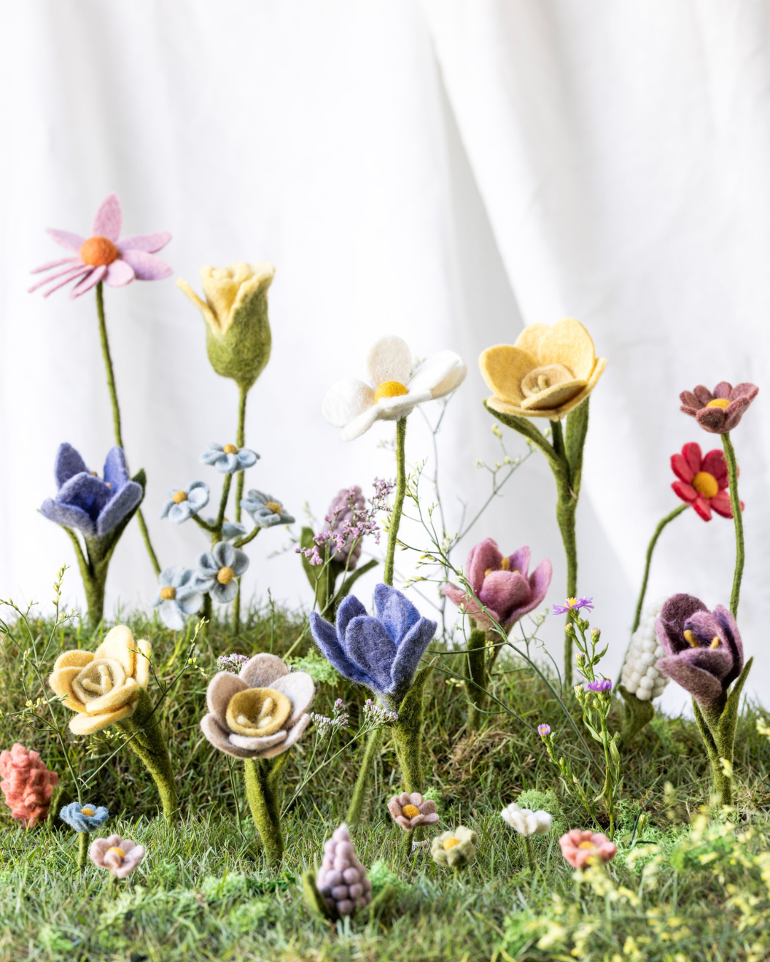 Frühlingsszene mit verschiedenen Filzblumen von Gry & Sif auf moosigem Untergrund, darunter Narzissen, Krokusse und weitere Blüten in zarten Farben vor hellem Stoffhintergrund.