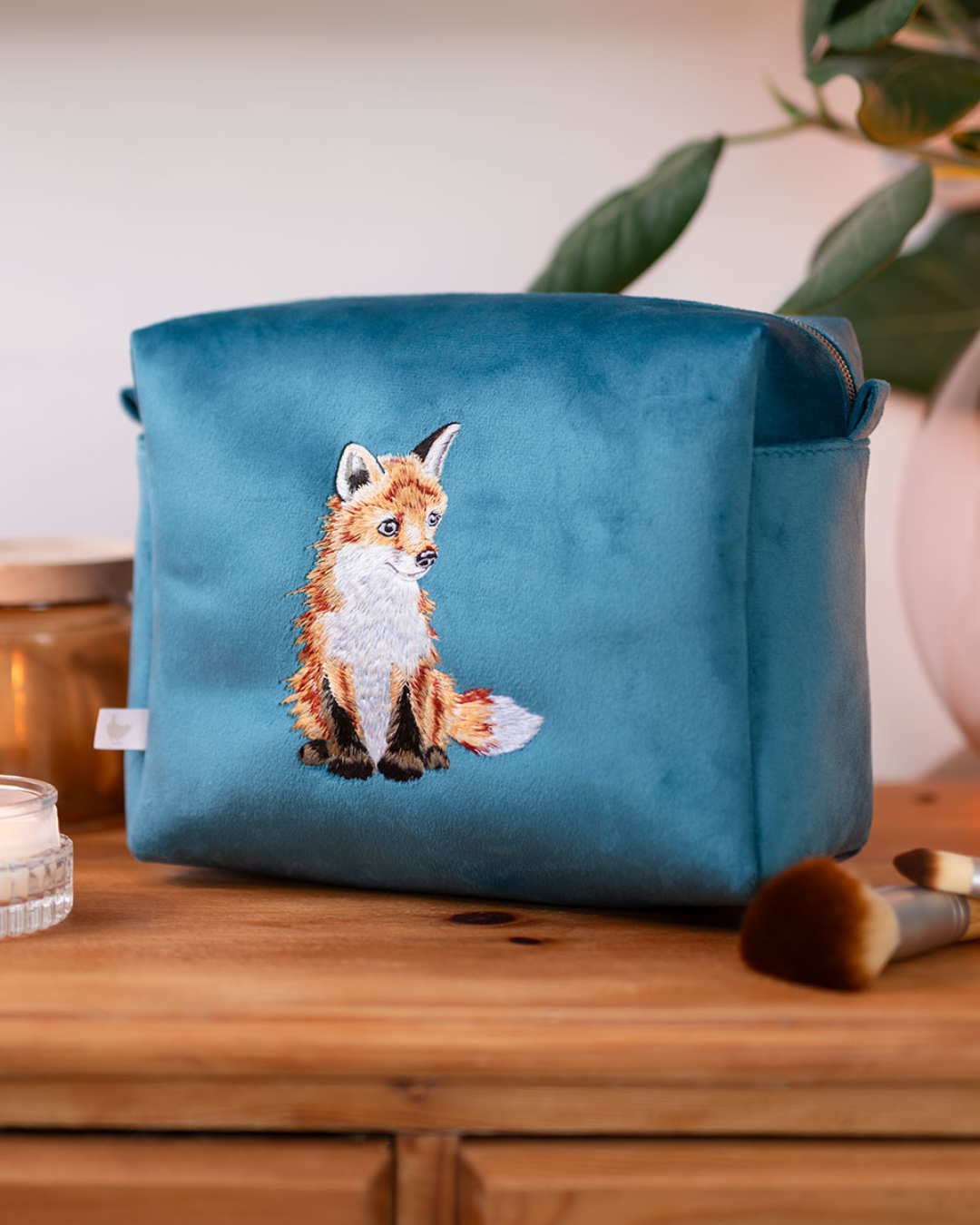 Große petrolfarbene Kosmetiktasche von Wrendale Designs mit gesticktem Fuchs-Motiv, platziert auf einer Holzablage mit Pinsel und Kerze.