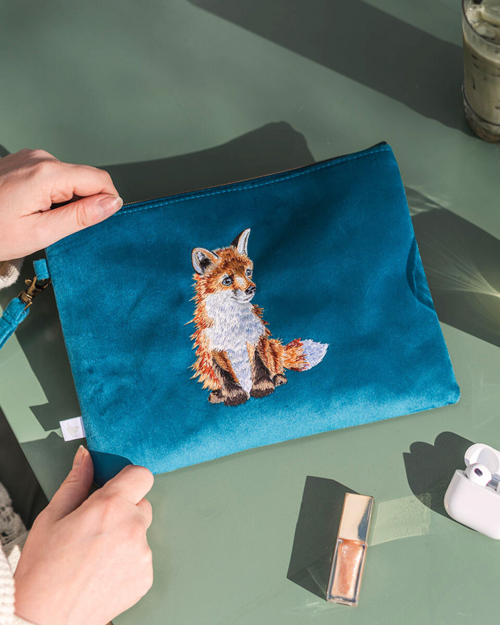 Petrolfarbener Reisebeutel von Wrendale Designs mit gesticktem Fuchs-Motiv, liegend auf einem Tisch mit Kosmetikartikeln und Kopfhörern.