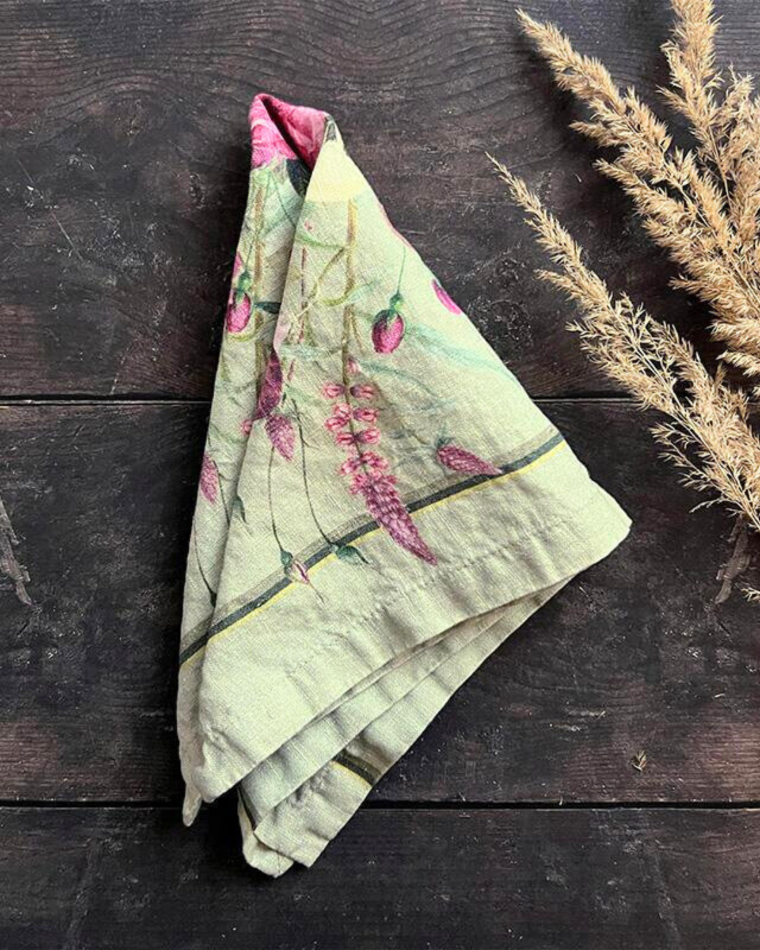 Eine einzelne, ausgebreitete Leinenserviette Blossom Garden von Koustrup & Co. auf dunklem Holzhintergrund, daneben eine natürliche Ähre als Dekoelement.