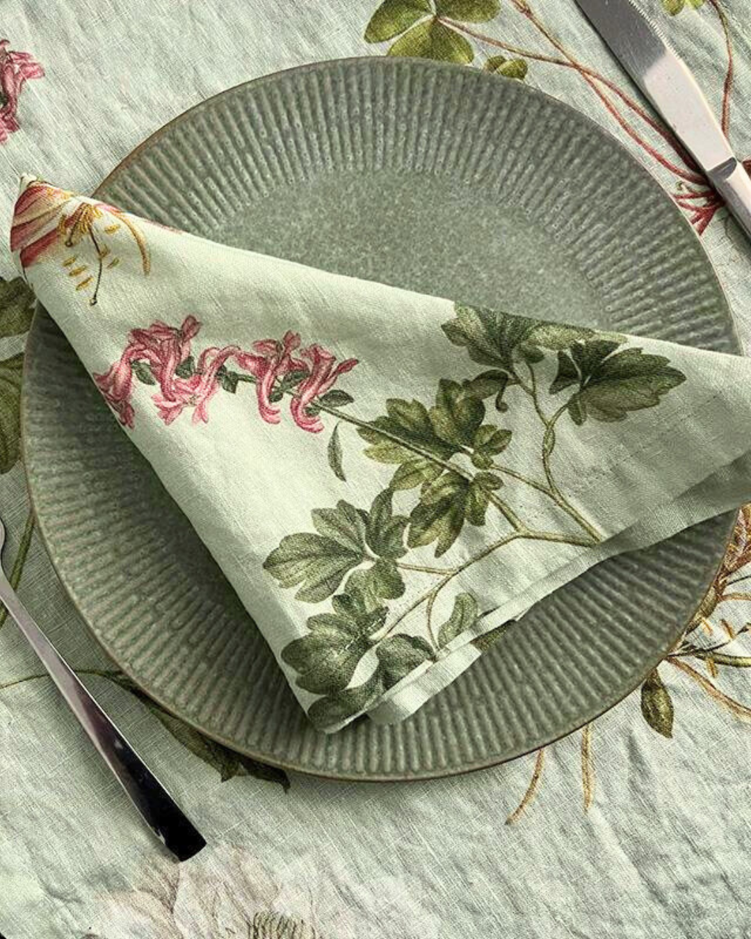 Gefaltete Stoffserviette mit floralen Details auf Grünem Teller welcher auf einem gedeckten farblich passenden Tisch liegt.