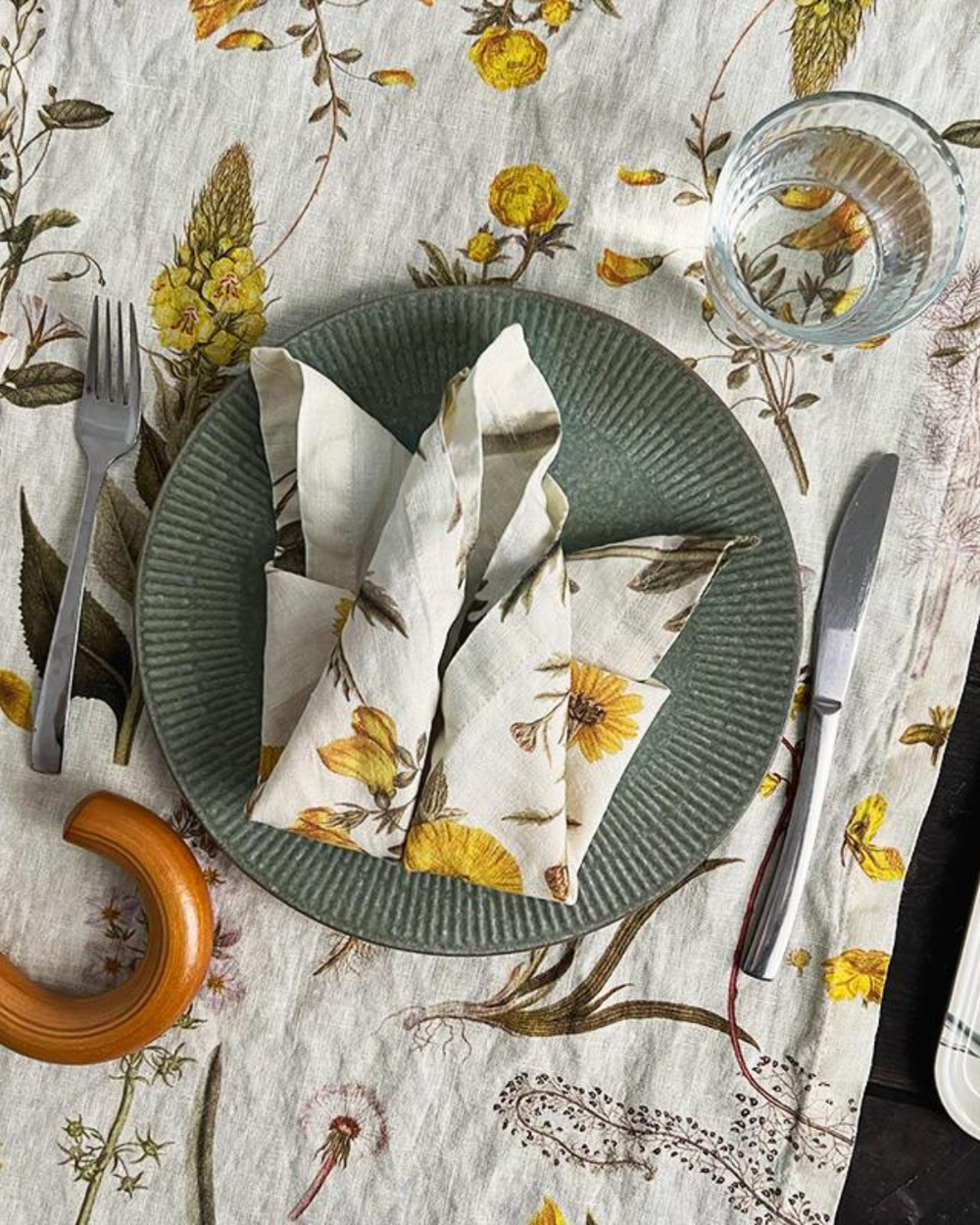Leinen Serviette "Flora Danica Atlas – gelb" von Koustrup & co gefaltet auf grünen Teller. Der Teller liegt auf einer ähnlich designten Tischdecke auf einem gedeckten Tisch.
