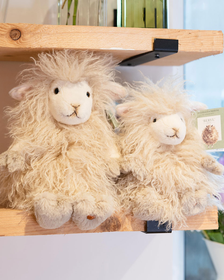 Schaf Kuscheliere Beryl in 2 Größen von Wrendale Designs sitzend auf einem Holzregal in einem dekorierten Zimmer. 
