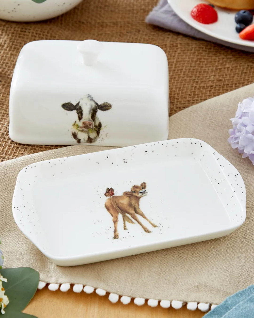 Geöffnete Kuh Butterdose "Royal Worchester" von Wrendale Designs auf bedeckten Tisch. Das Motiv auf dem Deckel zeigt eine schwarz weiße Kuh welche essend in die Kamera schaut. Auf der Unterseite ist eine braune Kuh mit einem Schmetterling zu sehen. Im Hintergrund sieht man ein Teller mit Früchten.