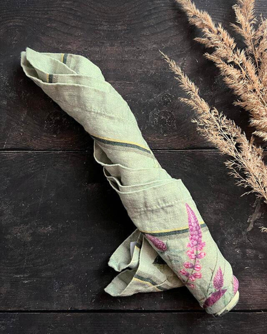 Eine gerollte Leinenserviette Blossom Garden von Koustrup & Co. auf dunklem Holztisch, daneben eine natürliche Ähre als Dekoration.
