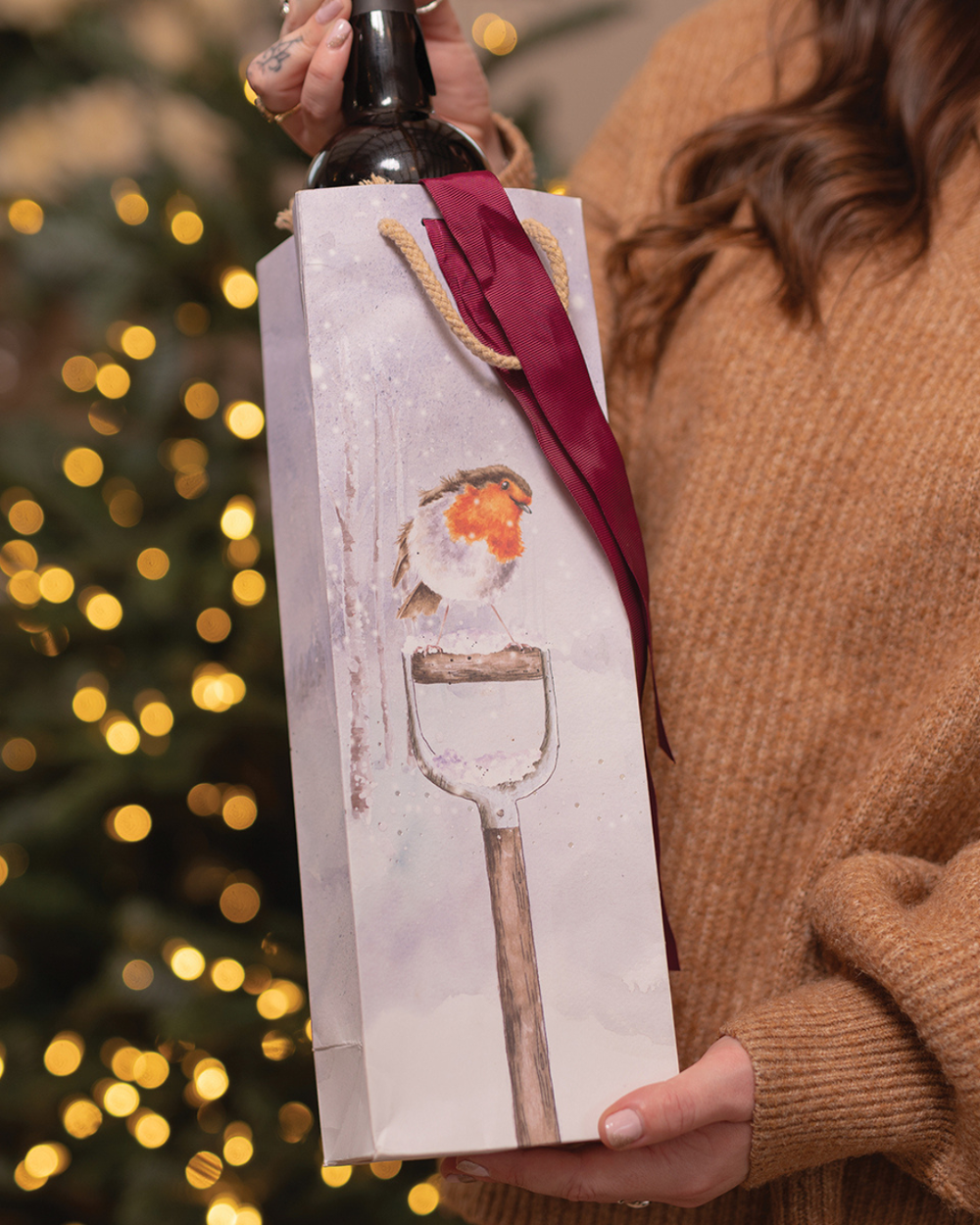 Person hält „A Little Red Robin“ Flaschen-Geschenktasche vor Weihnachtsbaum.