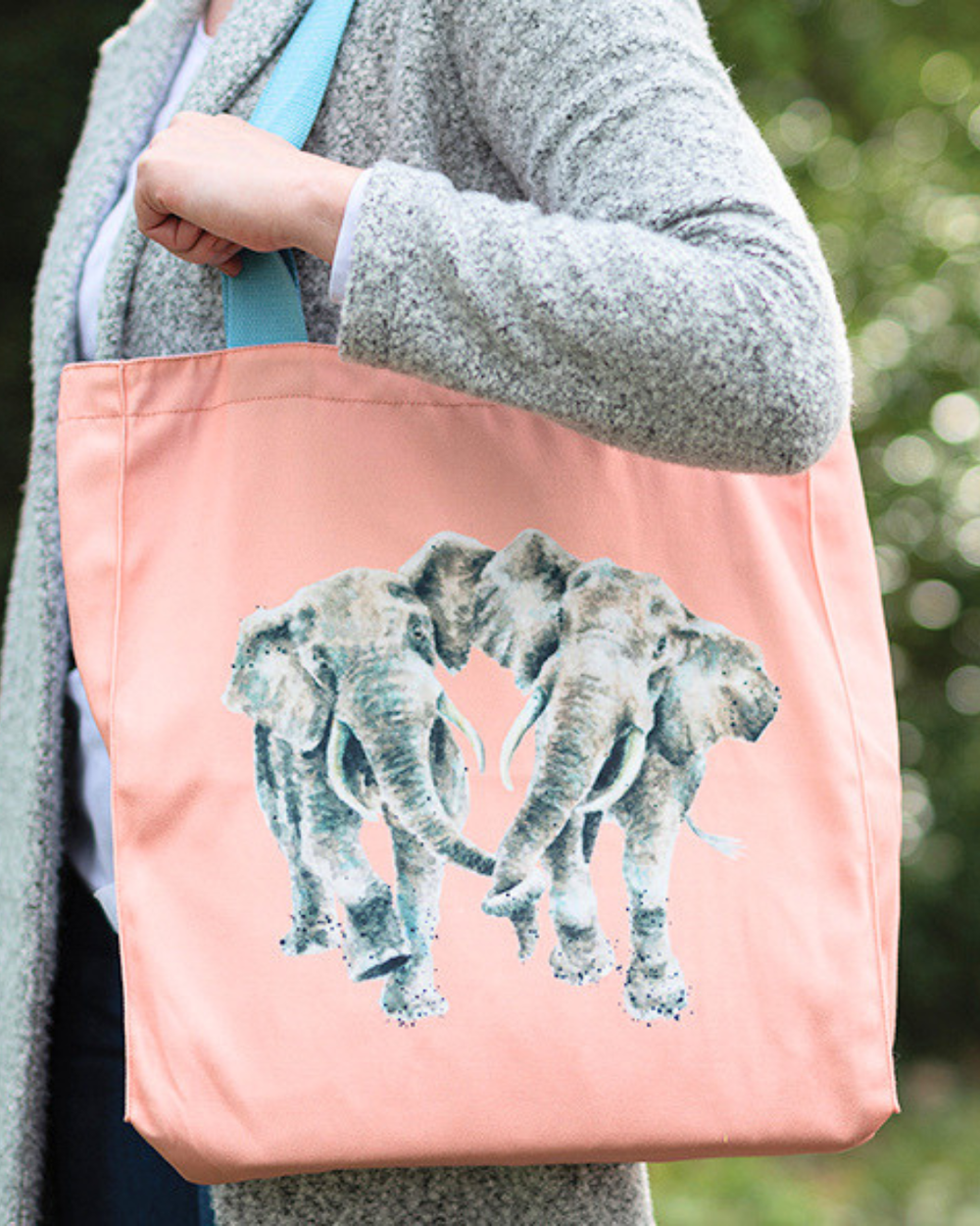 Pinke Elefanten Einkaufstasche von Wrendale Designs welche von einer Frau getragen wird. Das Motiv besteht aus 2 Elefanten die Händchen halten und der Griff ist hellblau.