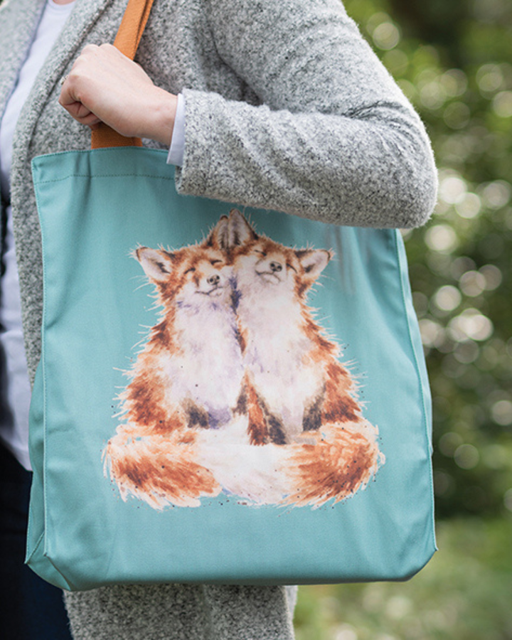 Blaue Fuchs-Einkaufstasche von Wrendale Designs getragen von einer Frau vor einem pflanzlichen Hintergrund. Zu sehen sind 2 Füchse welche liebevoll kuscheln. Der Griff der Tasche ist braun.