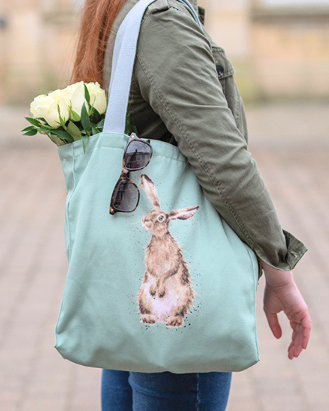 Hellgrüne Einkaufstasche von Wrendale Designs getragen von einer Frau. Aus der Tasche hängt eine Sonnenbrille und weiße Tulpen. Das Motiv ist ein neugierig schauender Hase. Der Griff der Tasche ist hellgrau.
