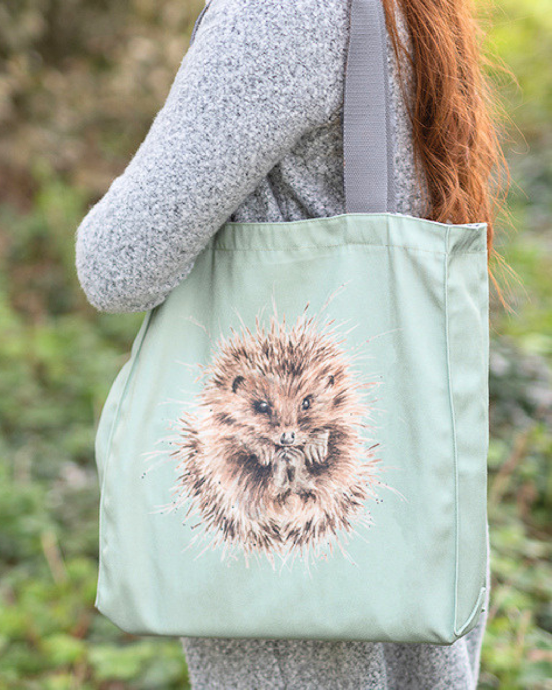 Hellgrüne Igel Einkaufstasche getragen von einer Frau im Freien. In der Mitte des Bilds ist ein niedlicher eingerollter Igel zu sehen, der Griff der Tasche ist grau. Im Hintergrund sind viele Pflanzen zu sehen.