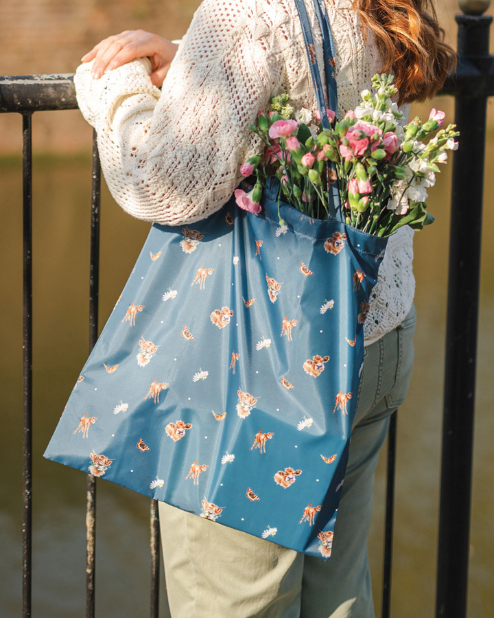 Dunkelblaue Einkaufstasche Farmyard Friends von Wrendale Designs in Benutzung. Zu sehen ist eine an einem Geländer stehende Frau, welche die Einkaufstasche über der Schulter trägt. Detailreiche Kälbchenillustrationen von Hannah Dale schmücken die Tasche. Im unteren Teil der Tasche zeichnen sich verstaute Einkäufe ab, während oben aus der Tasche heraus frische Schnittblumen in rosa und weiß zu sehen sind.