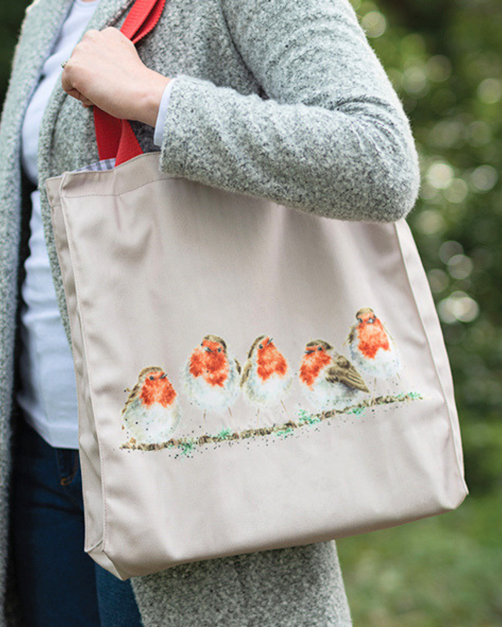 Rotkehlchen Einkaufstasche von Wrendale Designs. Zu sehen ist eine Frau mit blauer Jeans, weißem Oberteil und einem grauen Mantel welche die Hellgraue Rotkehlchen Tasche um ihre Schulter trägt.