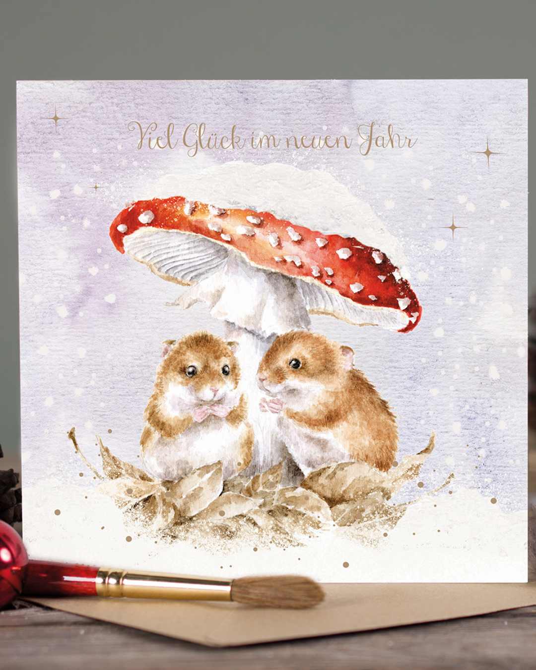 Die Doppelkarte Mushroom At The Inn illustriert von Hannah Dale für Wrendale Designs zeigt zwei niedliche braune Mäuse, die es sich unter einem roten, schneebedeckten Fliegenpilz auf etwas schneefreiem Laub bequem gemacht haben und etwas naschen. Der zarte Schriftzug Viel Glück im neuen Jahr steht in der oberen Bildmitte. Die Karte steht auf einem Holztisch. Im Vordergrund sind der braune Umschlag und ein rot-goldener Pinsel zu sehen.
