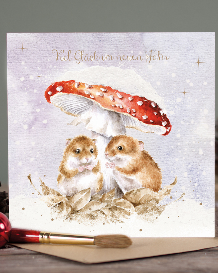 Die Doppelkarte Mushroom At The Inn illustriert von Hannah Dale für Wrendale Designs zeigt zwei niedliche braune Mäuse, die es sich unter einem roten, schneebedeckten Fliegenpilz auf etwas schneefreiem Laub bequem gemacht haben und etwas naschen. Der zarte Schriftzug Viel Glück im neuen Jahr steht in der oberen Bildmitte. Die Karte steht auf einem Holztisch. Im Vordergrund sind der braune Umschlag und ein rot-goldener Pinsel zu sehen.