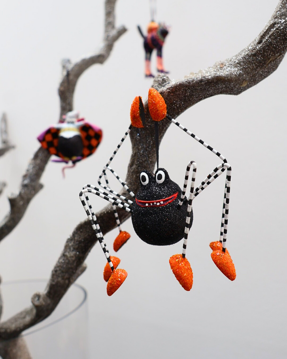 Krinkle-Spinne „Webster Spider“ von Patience Brewster hängt an einem dekorierten Ast, mit weiteren Halloween-Figuren im Hintergrund.