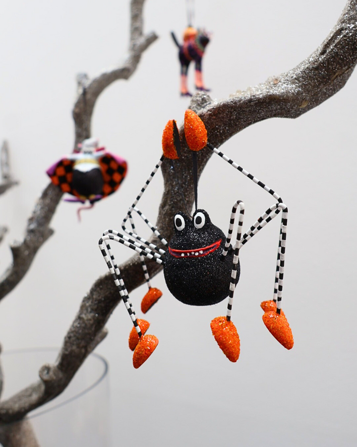 Krinkle-Spinne „Webster Spider“ von Patience Brewster hängt an einem dekorierten Ast, mit weiteren Halloween-Figuren im Hintergrund.