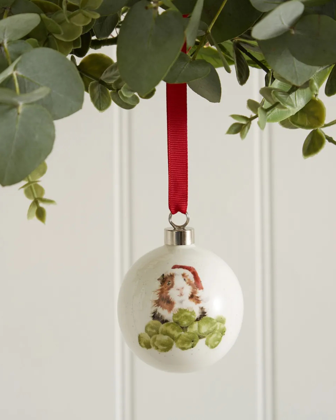 Weihnachtskugel mit Meerschweinchen-Motiv von Wrendale Designs aus Porzellan von Royal Worcester am roten Satinband hängend.