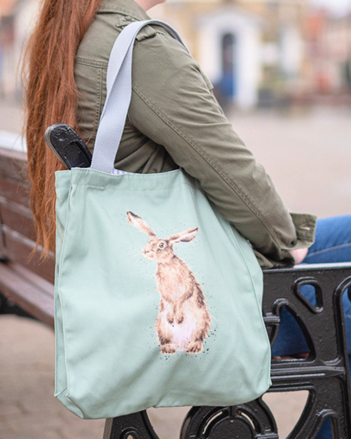 Hellgrüne Hasen-Einkaufstasche von Wrendale Designs getragen von einer Frau die auf einer Bank sitzt. Das Motiv ist ein neugierig schauender Hase und der Griff ist grau.