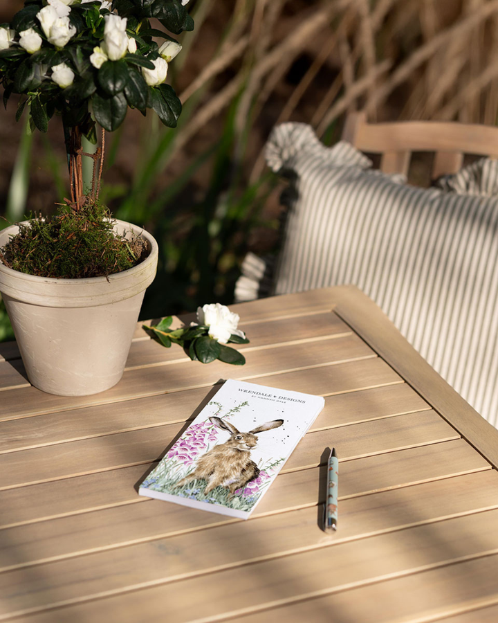 Hasen Notizblock "The WIld Garden" von Wrendale Designs auf Holztisch im Garten. Das Motiv ist ein Hase auf einer Blumenwiese welcher in die Kamera schaut. 