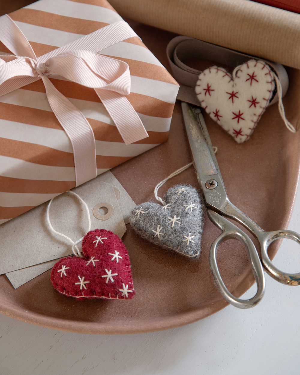 Herzanhänger aus Filz von Gry & Sif als festliche Geschenkdeko. Ein rotes, graues und weißes Herz mit gestickten Sternen neben einer Schere, Geschenkpapier und einem eingepackten Geschenk arrangiert.