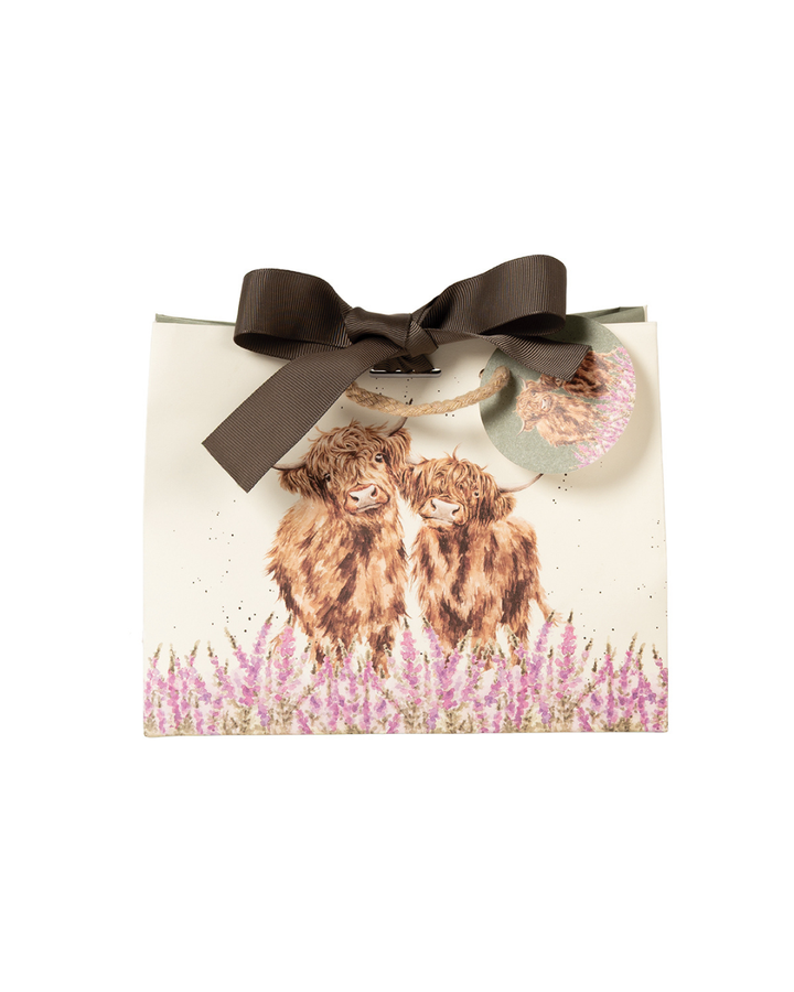 Die Geschenktasche zeigt zwei flauschige Hochlandrinder mit brauner Fellfarbe, die in einem Feld aus violett-rosanen Fingerhutblumen stehen. Im für Hannah Dale typischen natürlichen Stil blicken die beiden sanftmütig über die Blüten hinweg. Die Tasche ist am oberen Rand mit einer Schleife verschlossen. Ein Patch, welcher an der Verschlusskordel angebracht ist zeigt ebenfalls das Hochlandkuh-Motiv.