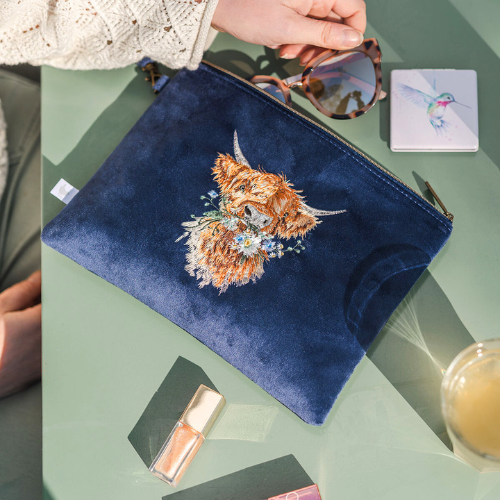 Dunkelblauer Reisebeutel von Wrendale Designs mit gesticktem Hochlandkuh-Motiv, liegend auf einem Tisch mit Kosmetikartikeln, einem Taschenspiegel und einer Sonnenbrille.