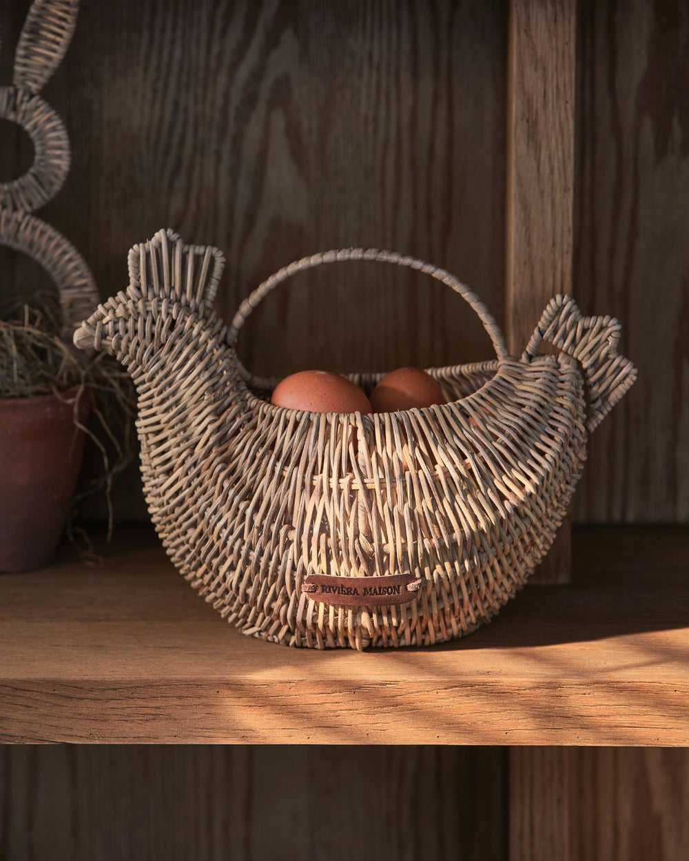 Rattan Korb in Form eines Huhnes von Rivièra Maison steht mit Eiern befüllt in einem Holzregal.