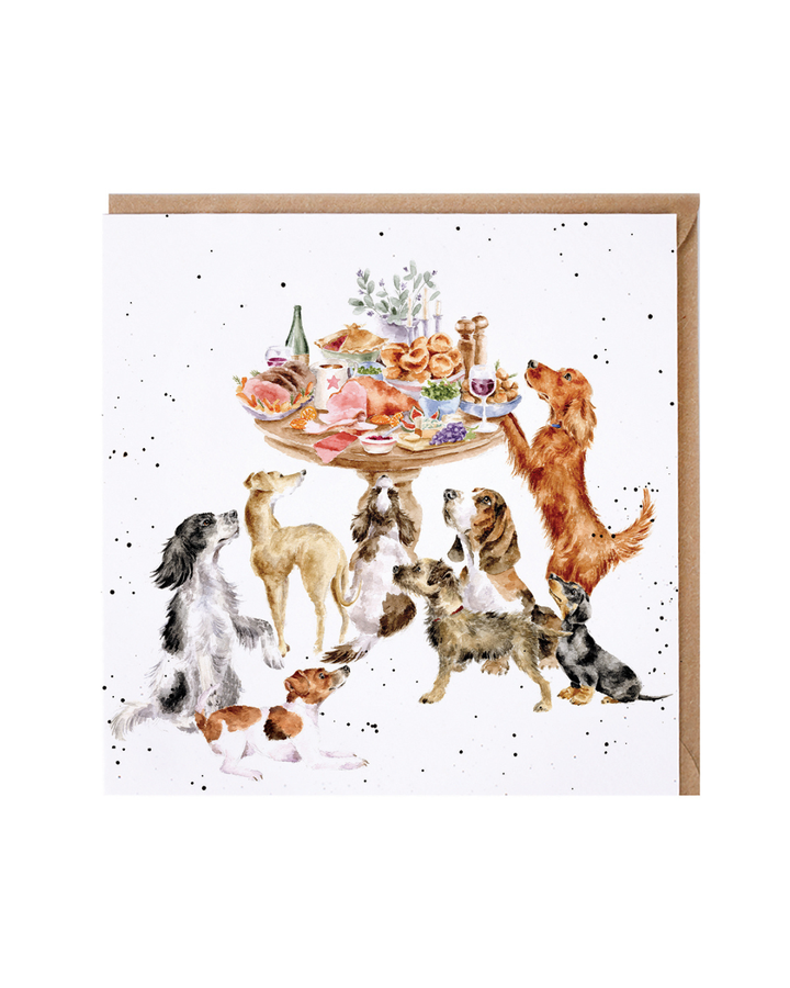 Hunde Grußkarte „Sunday Lunch“ von Wrendale Designs mit verschiedenen handillustrierten Hunderassen, die sich erwartungsvoll um einen gedeckten Tisch versammeln, weißer Hintergrund mit feinen Sprenkeln, brauner Umschlag im Hintergrund.