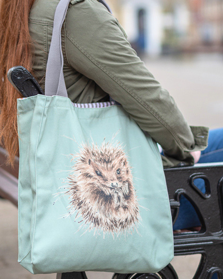 Hellgrüne Igel Einkaufstasche von Wrendale Designs getragen von einer sitzenden Frau im Freien. Das Motiv ist ein niedlicher eingerollter Igel. 