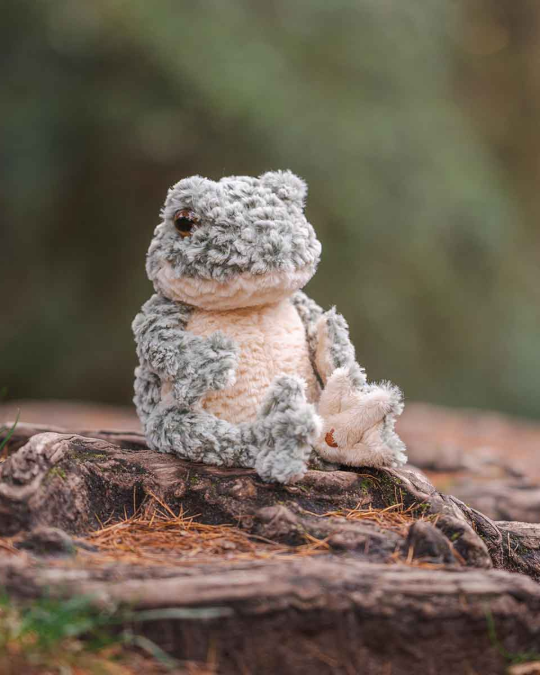 Seitlich fotografiertes Frosch Kuscheltier „Elliott“ von Wrendale Designs auf einem Baumstamm, mit Blick in die Waldszene.