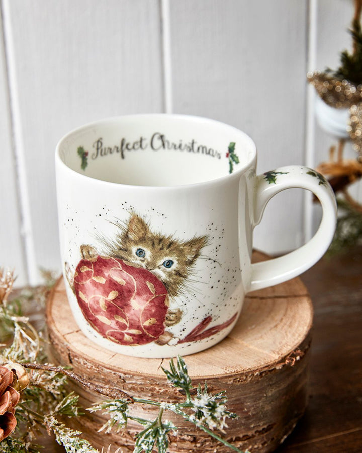 Das Motiv der Katzen Weihnachtstasse von Wrendale Designs zeigt eine Babykatze, die hinter einer Weihnachtsbaumkugel hervorschaut. Im Inneren der Tasse steht "Purrfect Christmas".
