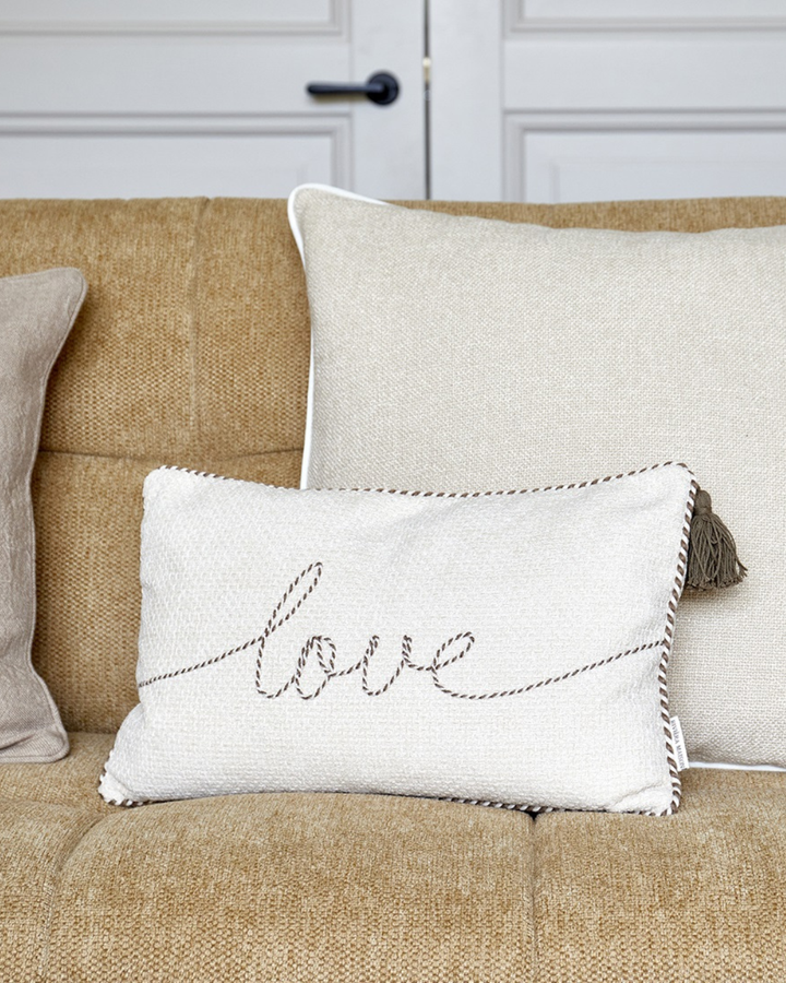 Kissenbezug „With Love Pillow Cover 50x30“ von Rivièra Maison auf einer Couch arrangiert, kombiniert mit größeren hellen Kissen vor einer hellen Tür.