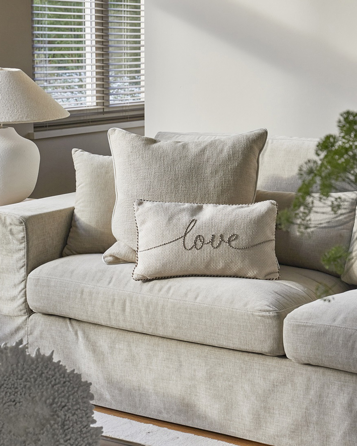 Kissenbezug „With Love Pillow Cover 50x30“ von Rivièra Maison auf einem Sofa in einem hellen Wohnzimmer, umgeben von weiteren Kissen, mit Fenster, Lampe und Tageslicht im Hintergrund.