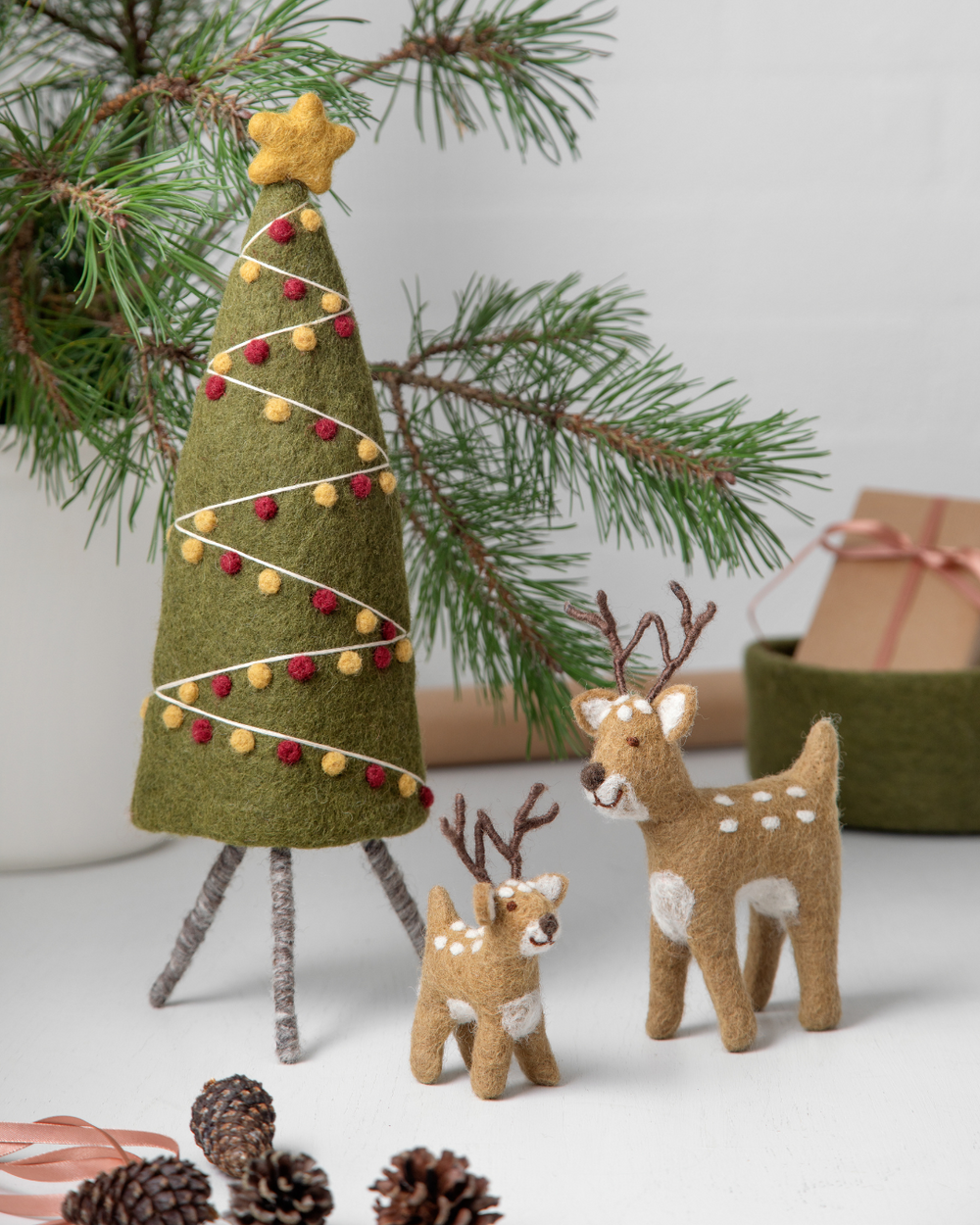 Weihnachtliche Szene mit Filzfiguren von Gry & Sif: grüner Filz-Tannenbaum mit gelben und roten Kugeln, daneben zwei handgefertigte Rentiere in Braun mit weißen Flecken und Geweihen, dekoriert mit Tannenzweigen und Kiefernzapfen.
