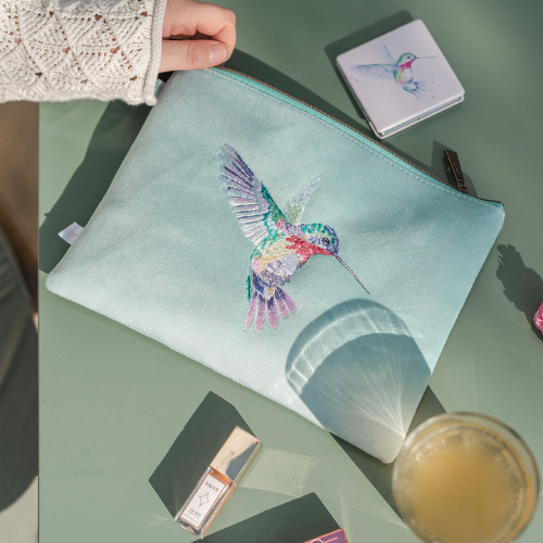 Türkisfarbener Reisebeutel von Wrendale Designs mit gesticktem Kolibri-Motiv, liegend auf einem Tisch mit Kosmetikartikeln.
