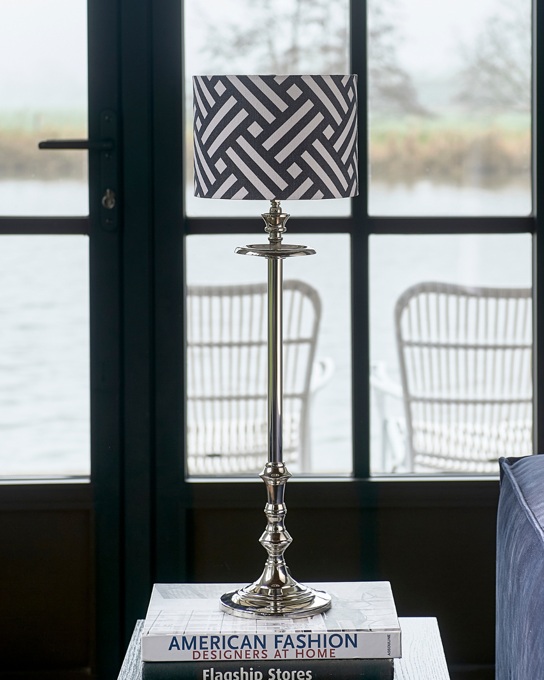 Lampenfuß L'Hotel Lampbase von Rivièra Maison mit grafisch gemustertem Lampenschirm auf Stapel Büchern, platziert vor großer Fensterfront mit Blick nach draußen.