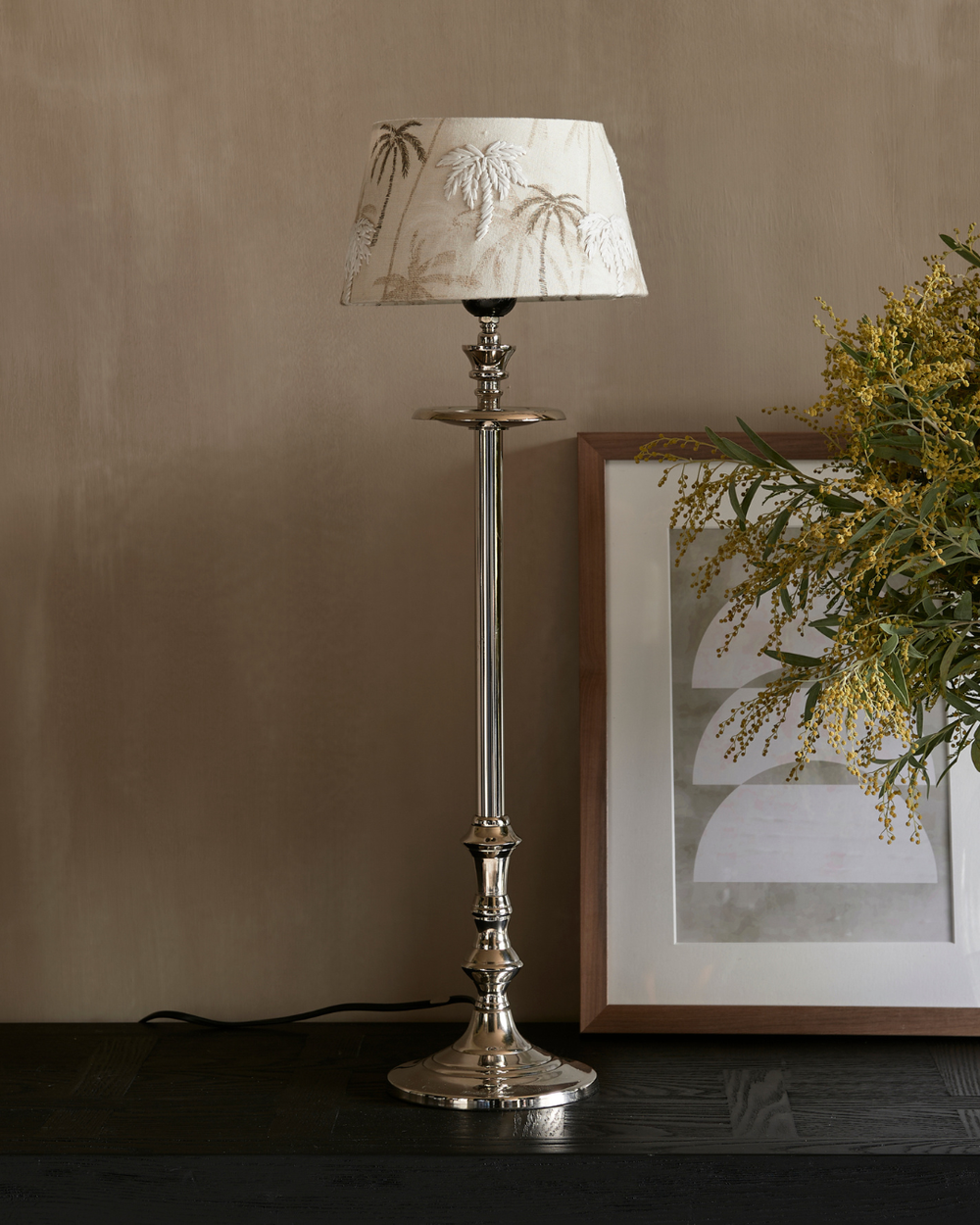 Lampenfuß L'Hotel Lampbase von Rivièra Maison aus glänzendem silberfarbenem Metall mit beigem Lampenschirm, dekoriert auf dunklem Sideboard vor ruhiger Wand.