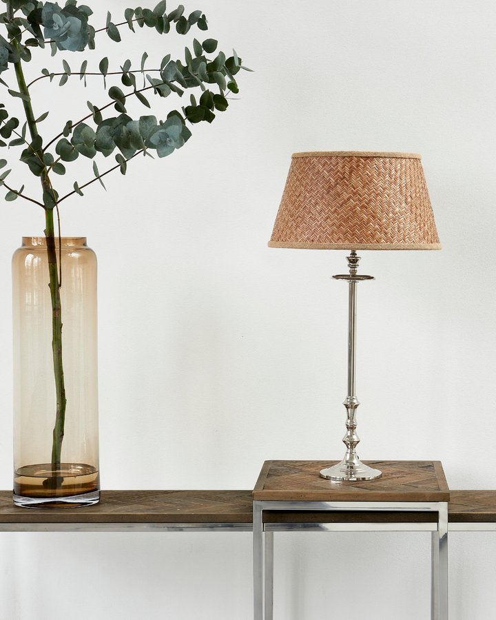 Lampenfuß L'Hotel Lampbase von Rivièra Maison mit geflochtenem Lampenschirm auf Holzkonsole, neben transparenter Glasvase mit Zweigen vor heller Wand.