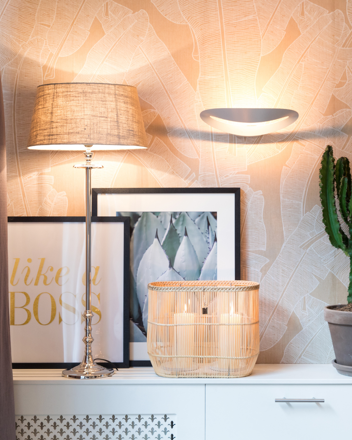 Eingeschalteter Lampenfuß L'Hotel Lampbase von Rivièra Maison mit Stofflampenschirm in warmem Licht, arrangiert zwischen Bilderrahmen und Pflanze auf weißem Möbelstück.