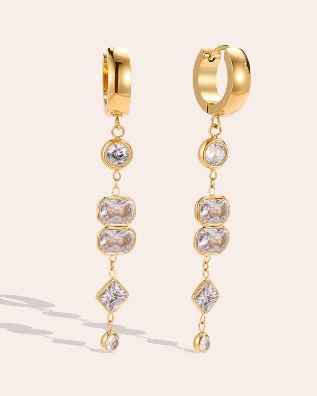 Lange Ohrringe mit weißen Zirkoniasteinen von Dottilove vor beigen Hintergrund. Die Ohrringe sind gold farbig am unteren ende ist eine Kette befestigt an denen die weißen Steine hängen. 